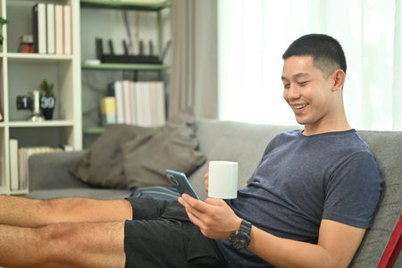 Smiling man typing massage, checking social media on mobile phone while leaning on couch at home.の写真素材