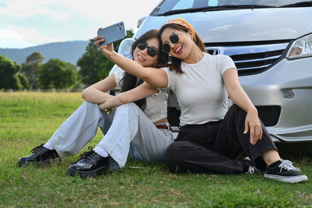 Cheerful female friends taking a break from driving siting in front of car on meadow and taking...