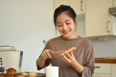Happy attractive young woman housewife preparing breakfast spreading strawberry jam on slice of bread.の写真素材