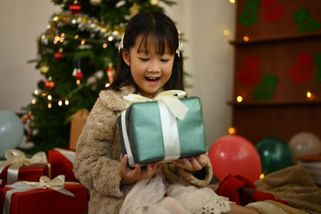 Excited girl holding Christmas gift box with a beautifully decorated Christmas tree on background in a cozy living room.の写真素材