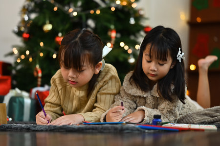 Two cute little girls making greeting card for New Year and Christmas while lying on floor in decorated living room.の写真素材