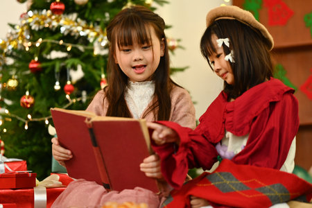 Two happy beautiful little girl reading a storybook together in a cozy living room on Christmas eve.の写真素材