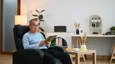 Calm senior man in casual clothes resting on armchair and reading favorite literature.