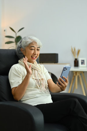 Beautiful gray haired mature woman waving hand while talking online on smartphone.の写真素材