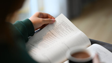 Closeup view of young woman drinking coffee and reading book on couch at home.
