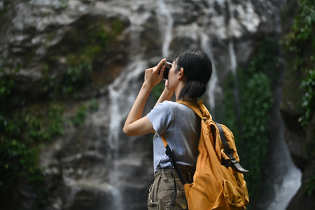 Young woman backpacker standing in front of waterfall and taking photos with vintage retro camera.の写真素材