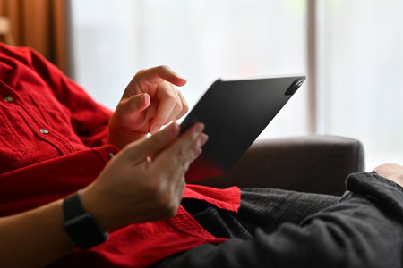 Cropped shot of man in red shirt sitting on sofa and checking social media on digital tablet.