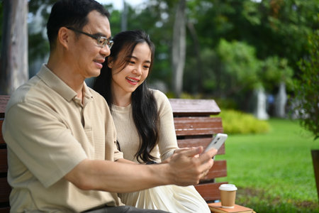 Happy middle age man and adult daughter watching funny video on smart phone while sitting on bench in the park.の写真素材