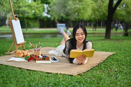 Carefree young woman lying on mat and reading interesting book at beautiful green park.の写真素材