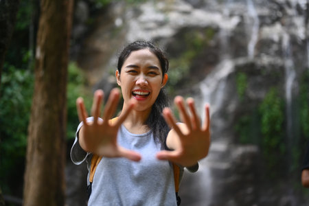 Portrait of happy young woman tourist with backpack standing in front of the tropical waterfall.の写真素材