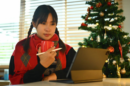 Pretty young woman wearing warm sweater drinking coffee and reading email on digital tablet.