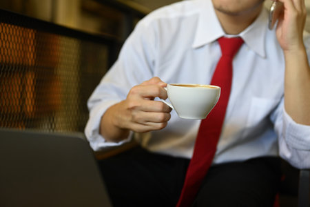 Businessman in white shirt holding cup of coffee and talking on mobile phone at cafe.の写真素材