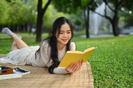 Relaxed young woman lying down on mat and reading interesting book at green park.