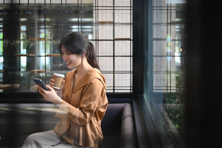 Attractive young businesswoman with takeaway coffee using mobile phone at corporate office break out area.の写真素材