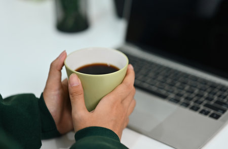 Close up of woman hands holding cup of hot tea and using laptop at office desk.の写真素材