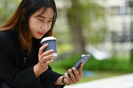 Happy young Asian woman drinking coffee and reading message on mobile phone.の写真素材
