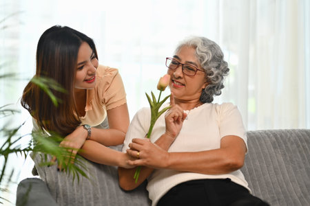 Adult daughter standing behind couch and giving flowers to older mother. Family bonding concept.