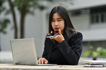Beautiful woman in formal clothes talking on mobile phone and using laptop outdoor.の写真素材