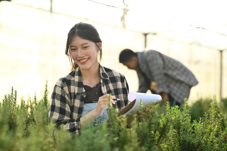 Attractive female farmer with clipboard doing quality check of organic rosemary in greenhouse.