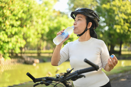 Active senior woman drinking water from a bottle after workout against blurred green park backgroundの写真素材