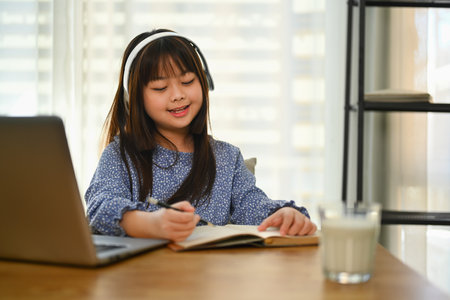 Smiling Asian schoolgirl in headphone studying online on laptop, doing homework at dinning tableの写真素材