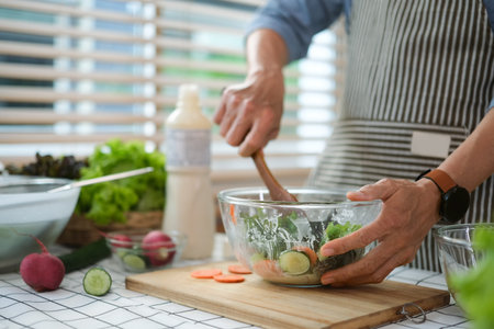 Middle age man standing in kitchen table making vegetarian salad with fresh farm vegetablesの写真素材