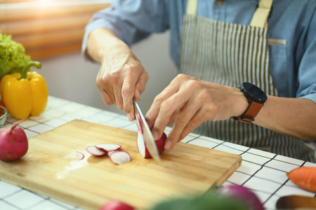 Senior man wearing apron cooking healthy food at home, slicing fresh radishes on cutting boardの写真素材