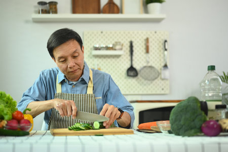 Middle age man cutting cucumber on board preparing wonderful fresh vegan salad in the kitchenの写真素材
