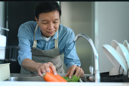 Smiling retired man washing vegetables in kitchen sink before cooking healthy foodの写真素材