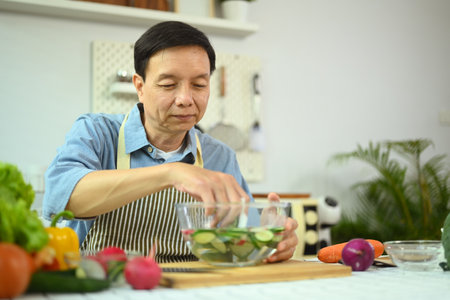 Smiling retired man making fresh vegetable salad, enjoying cooking alone at homeの写真素材