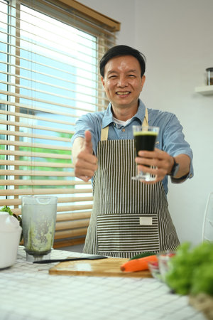Cheerful middle age man holding glass of green smoothie and showing thumbs upの写真素材