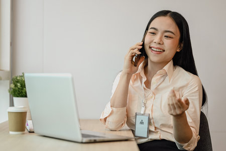 Attractive Asian female employee talking on mobile phone and using laptop at desk.の写真素材