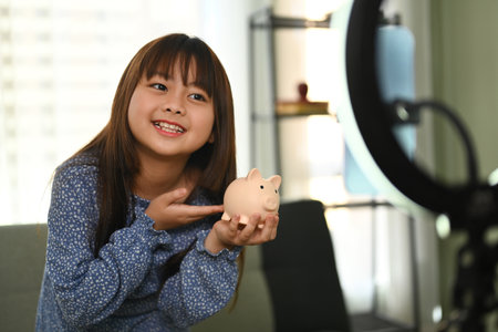 Pretty asian girl holding piggybank in front of camera while recording video on smartphone. Saving money for future concept.の写真素材