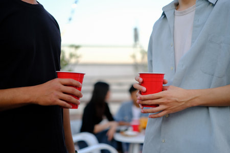 Two young male friends holding plastic glasses with beer at outdoor rooftop party.の写真素材