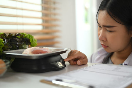 Female dietitian weighing raw chicken breast on a scale while working on a diet plan at clinic.の写真素材