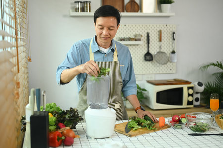 Happy middle age man making vegetable smoothies with blender in kitchen. Healthy lifestyle concept.の写真素材