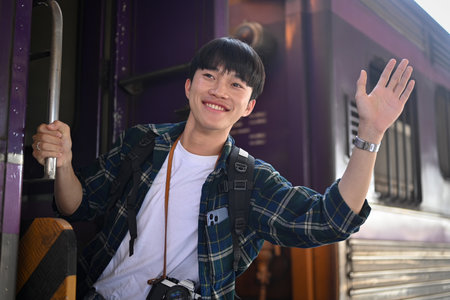 Happy young Asian man waving hand to friend while standing in train door before leaving.の写真素材