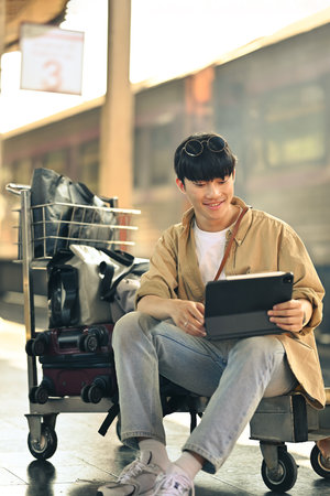 Smiling young man sitting on luggage cart and using digital tablet, waiting for the train.の写真素材