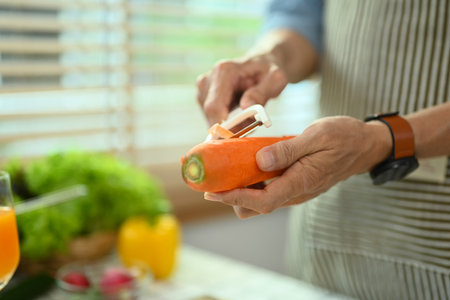 Cropped shot senior man in apron peeling carrot, preparing healthy salad in kitchen.の写真素材