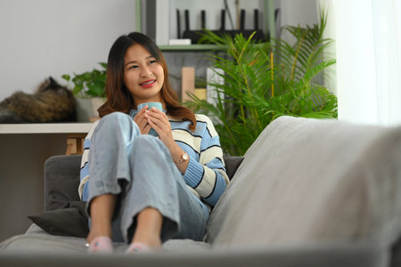 Attractive young woman sitting on sofa at home with cup of coffee and looking aside with smile.の写真素材