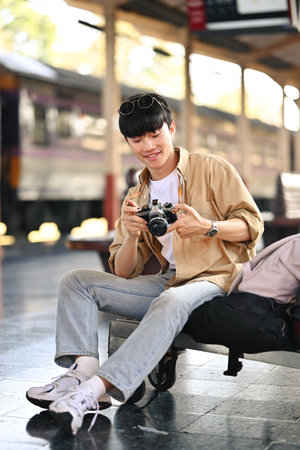 Full length young man tourist checking picture on camera while sitting at railway station.の写真素材
