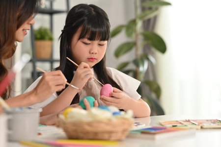 Cute little girl painting eggs with her mother, preparing decorations for easter celebration at...