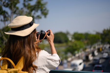Young woman wearing straw hat and backpack taking photo with retro camera on river bridge.の写真素材