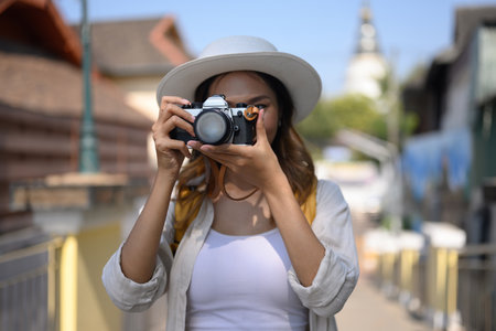 Young beautiful tourist with backpack taking photos with retro film camera at local city street in...
