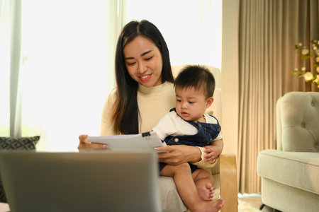 Happy Asian mother with baby boy using laptop for online work in living room at home.
