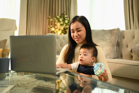 Young asian mother holding her baby boy on hands while working with laptop at home.の写真素材