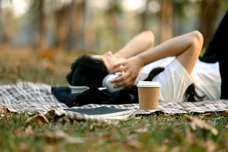 A paper cup of coffee on checkered blanket near relaxed young woman listening to music with...