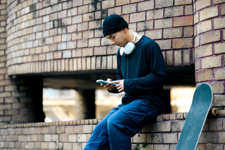 Handsome Asian man in stylish clothes using mobile phone while sitting with skateboard at city street.の写真素材