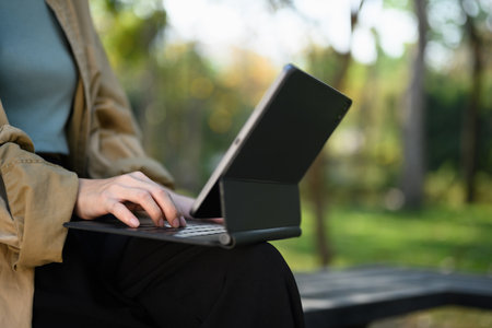 Cropped shot young lady freelancer working with digital tablet, sitting on wooden bench in park.の写真素材