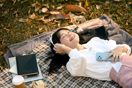 Happy young Asian girl in headphones lying on picnic blanket with closed eyes.
